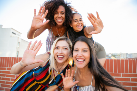 Four women with different looks and styles huddle close and pose together, waving at the camera.